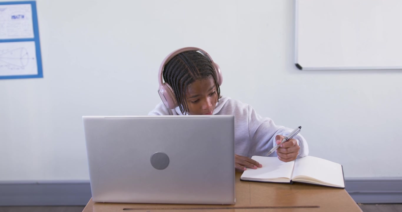 Teenage African American male student wearing pink headphones, studying on laptop and writing notes