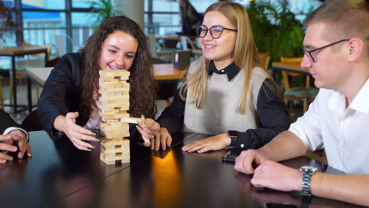Cheerful happy group of young people play board game. Woman with long curly hair pulling brick from tower and other people watching her.