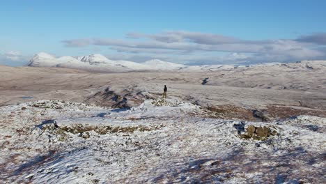 Vista-Aérea-De-Paralaje-De-Un-Hombre-Parado-En-La-Colina-De-Ullapool-Cubierta-De-Nieve-En-Escocia