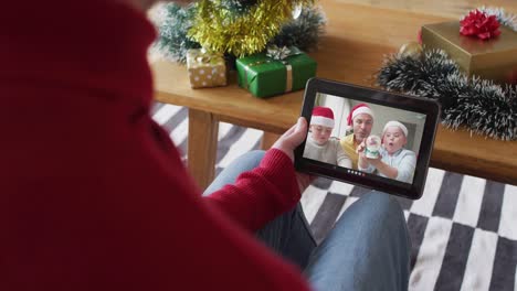 Hombre-Caucásico-Saludando-Y-Usando-Una-Tableta-Para-Una-Videollamada-Navideña-Con-Una-Familia-Sonriente-En-La-Pantalla