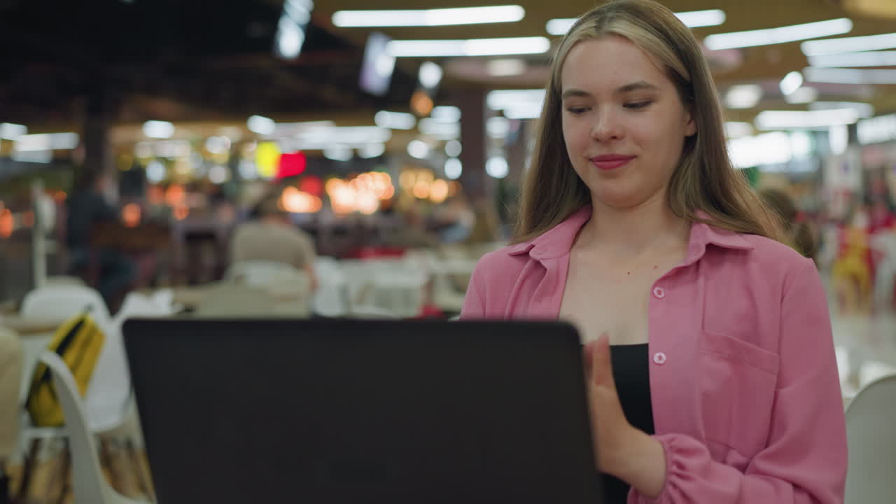mujer blanca en vestido rosa se sienta frente a su portátil, sonriendo con anticipación mientras mueve la cabeza de izquierda a derecha, en un restaurante ocupado y bien iluminado, con una suave iluminación bokeh en el fondo