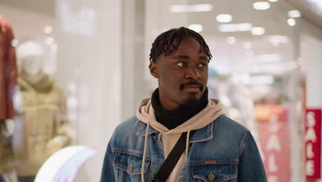 young black man in denim jacket and hoodie looking upward in shopping mall, browsing store displays, modern glass storefronts, sale signage, thoughtful expression, lifestyle