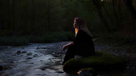woman sitting by a tranquil forest stream at dusk