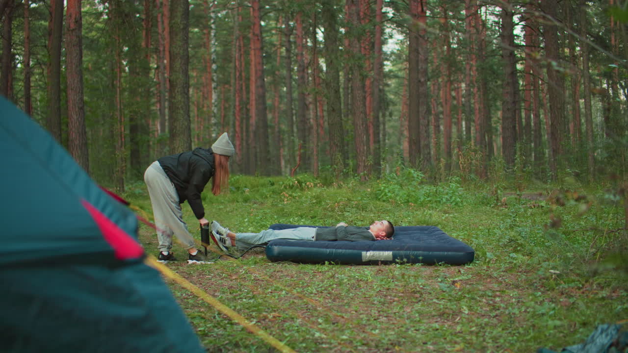 man lies on air bed resting while woman pumps air into it with pitched tent behind in dense forest clearing, highlighting camping teamwork, relaxation, and nature immersion