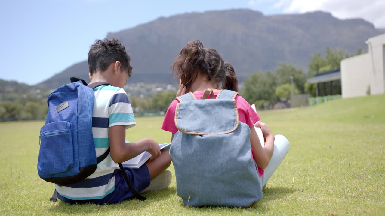 Biracial boy and girl sit on grass with backpacks at school, facing a mountain