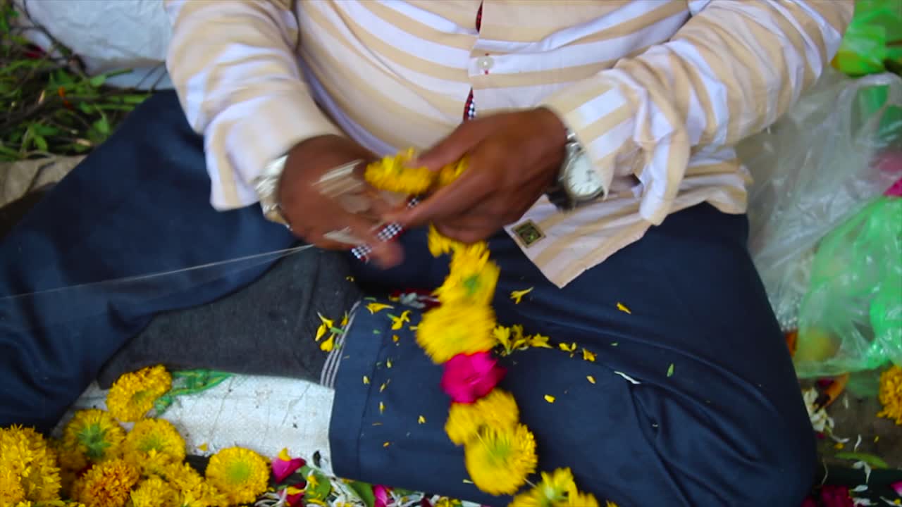 Close-up shot of man making marigold and rose garland by hand. Used for weddings, temples and festivals. Great for culture, craft and devotion-related footage.