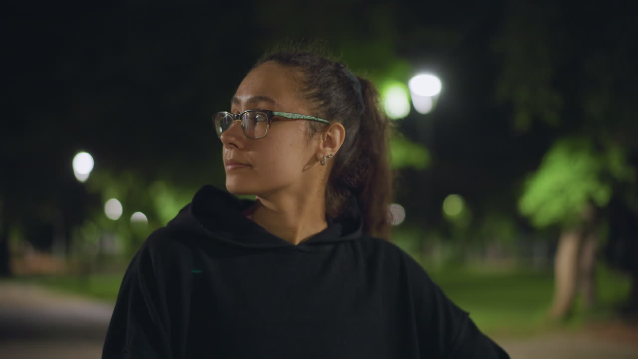 Primer plano de una mujer joven en un parque de noche, bokeh suave de una farola detrás de los árboles, gafas y expresión de perfil serena, ambiente introspectivo con luz fría al atardecer y sutil vegetación urbana de fondo