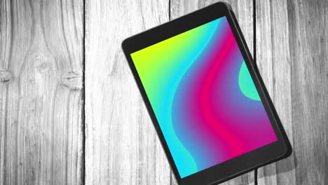 Animation-of-tablet-with-colorful-moving-shapes-on-screen-on-wooden-background