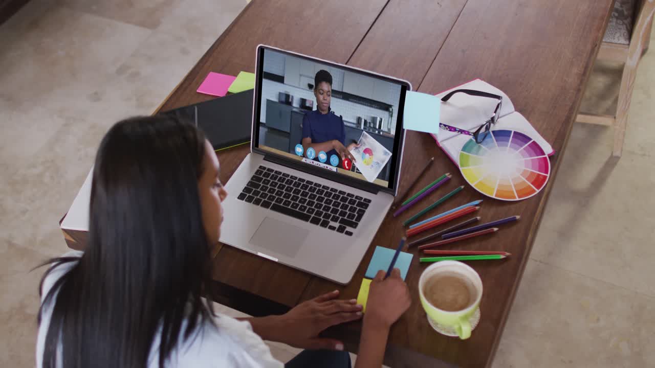 mujer caucásica usando una computadora portátil en videollamada con una colega y tomando notas