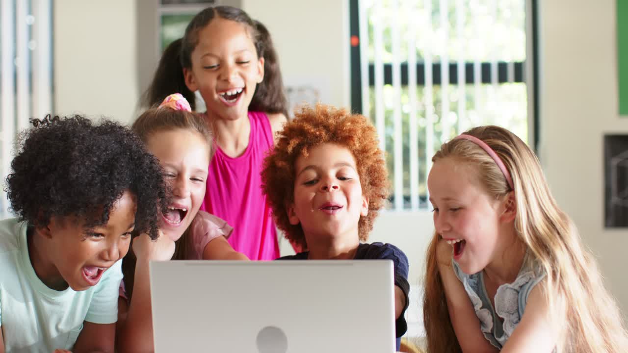 Diverse children friends leaning around laptop at classroom table, reacting to clip and laughing