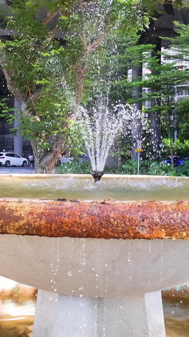 Paneo de cámara diurno sobre una fuente exterior moderna, rociando agua entre vegetación y edificios de la ciudad.