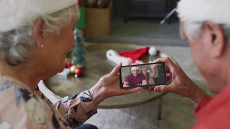 pareja caucásica mayor sonriente usando un teléfono inteligente para una videollamada de navidad con la pareja en la pantalla