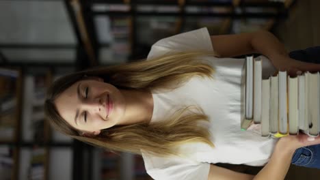 Student-girl-walks-through-library-with-stack-of-books-in-hands,-shelves-with-book,-front-view.-Young-woman-hold-books-at-hands