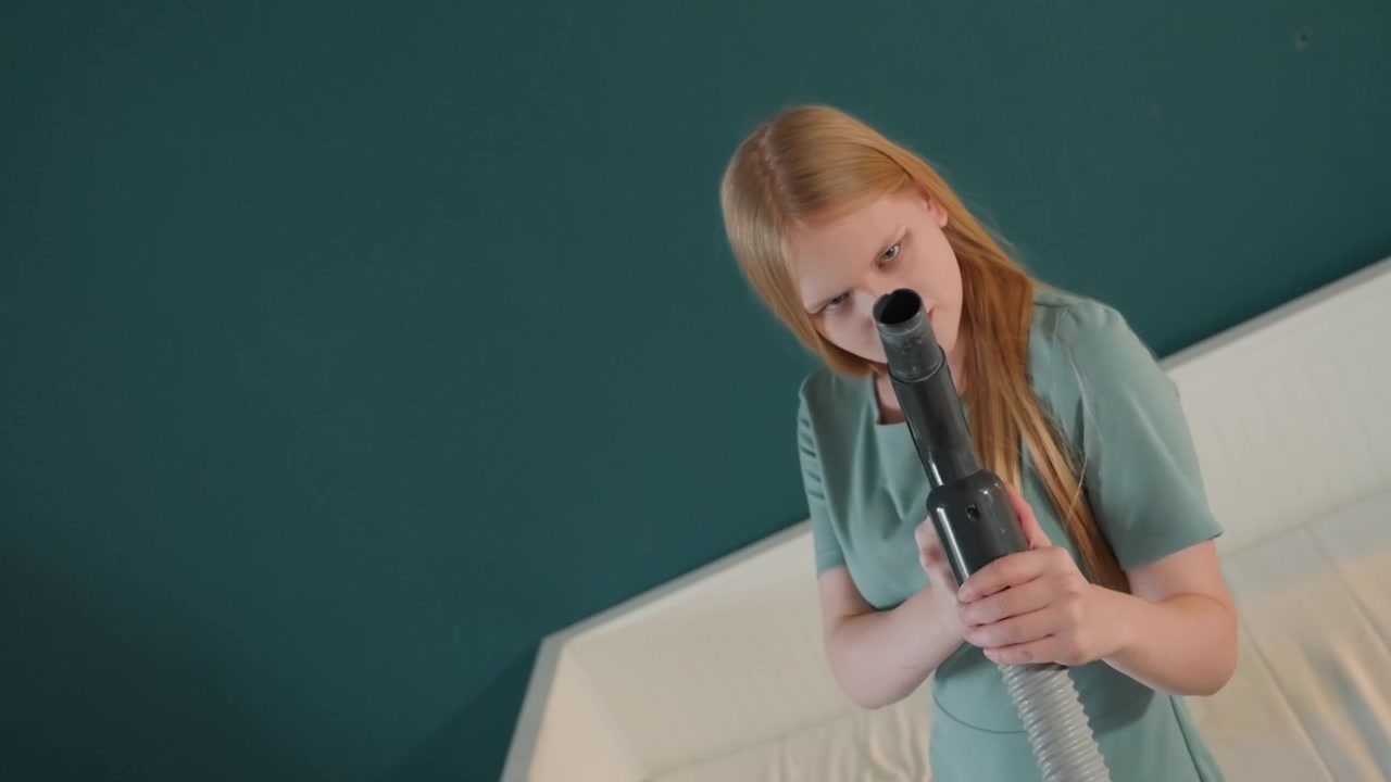 Donna caucasica che aspira il letto della camera da letto, movimenti lenti e metodici, sfondo di parete color verde acqua, struttura del letto e biancheria bianche, bocchetta dell'aspirapolvere concentrata sulle cuciture del materasso, luce del mattino, routine ordinata, ambiente domestico tranquillo