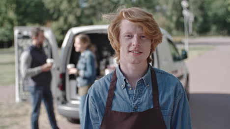 coffee truck redhead worker looking at camera smiling, a group of people talk and drink coffee in the background