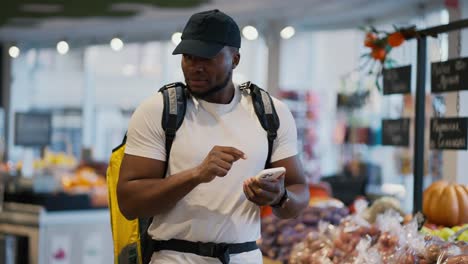 Un-Repartidor-De-Piel-Negra-Con-Camiseta-Blanca-Selecciona-Y-Busca-Los-Productos-Necesarios-Para-La-Entrega-De-Comida-Y-Sostiene-Una-Gran-Bolsa-Amarilla-Sobre-Sus-Hombros-En-Un-Supermercado-Moderno.