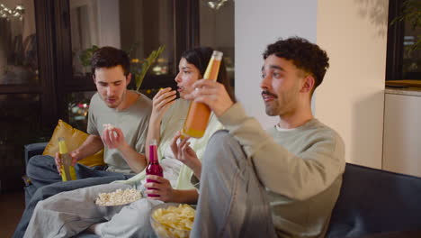 vista lateral de tres amigos viendo una película interesante en la televisión sentados en el sofá, comiendo palomitas de maíz y papas fritas y bebiendo refrescos 1