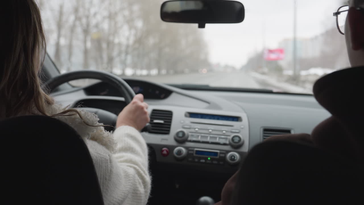 inside view of woman in white sweater driving on open road while wiper cleans moist windshield, passing vehicles and road signs visible ahead, trees and overcast sky