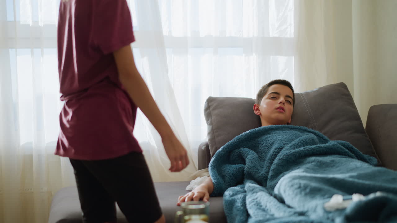 Boy lying on couch under a blue blanket, surrounded by tissues, while his sister gently covers him, natural light filters through the window creating a cozy, caring atmosphere