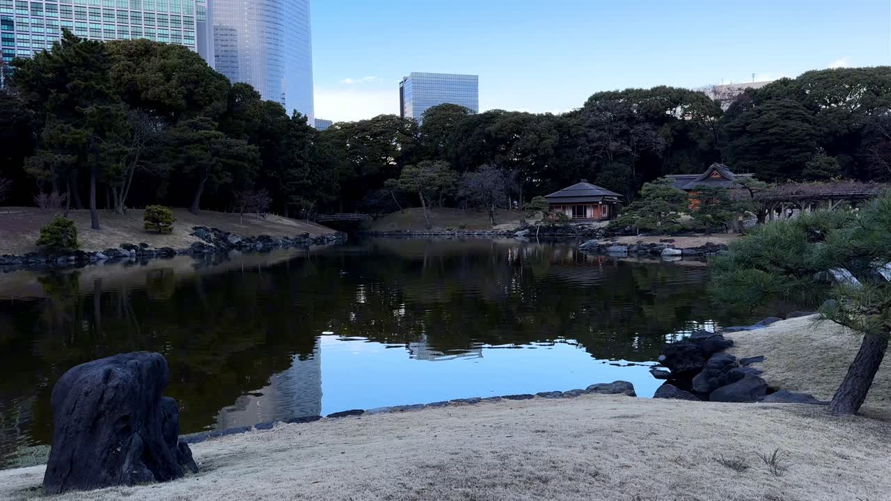 A serene pond surrounded by trees, traditional buildings, and city skyscrapers nearby