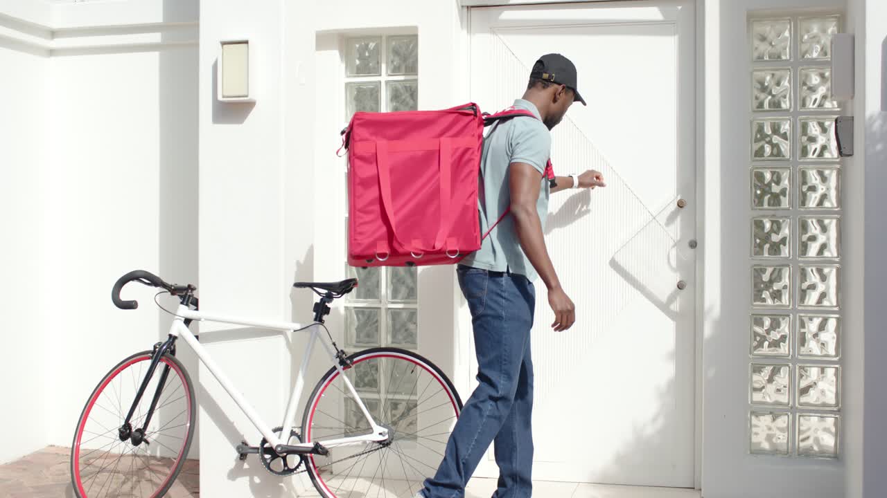 Courier arriving at stucco house parking bike adjusting red delivery backpack and pressing intercom