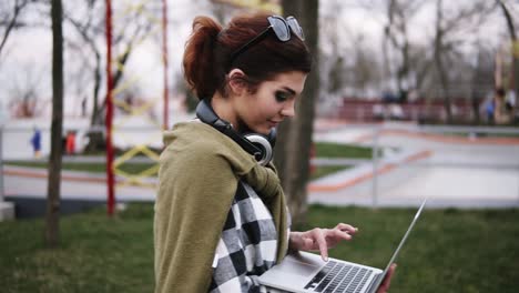 una joven con gafas de sol en la cabeza y una camisa a cuadros camina por el parque, su computadora portátil está en su mano. ella va y charla al mismo tiempo, escribiendo texto. vista lateral