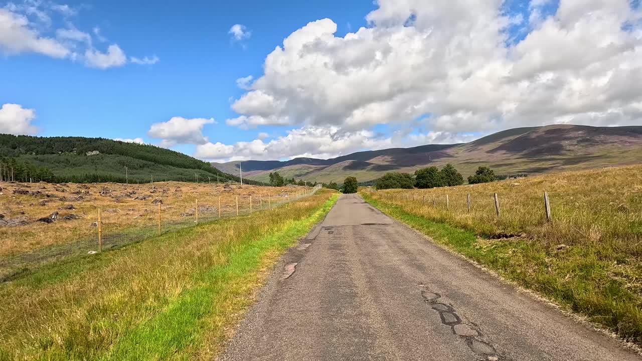 A vehicle travels a narrow country road through scenic Scottish hills under bright daylight, with gentle camera movement and expansive landscape views