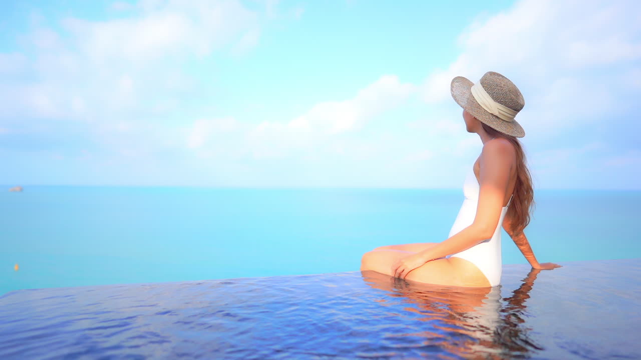 Asiatische Frau in weißem Badeanzug und breitem Sonnenhut sitzt mit den Füßen zusammen im Wasser am Rand des hohen Infinity-Pools auf dem Dach vor einem Hintergrund aus Himmel und Meereslandschaft.