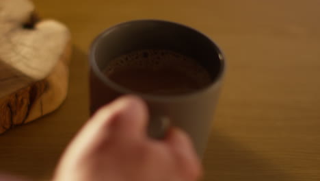 close up of person picking up steaming cup of hot chocolate drink on table