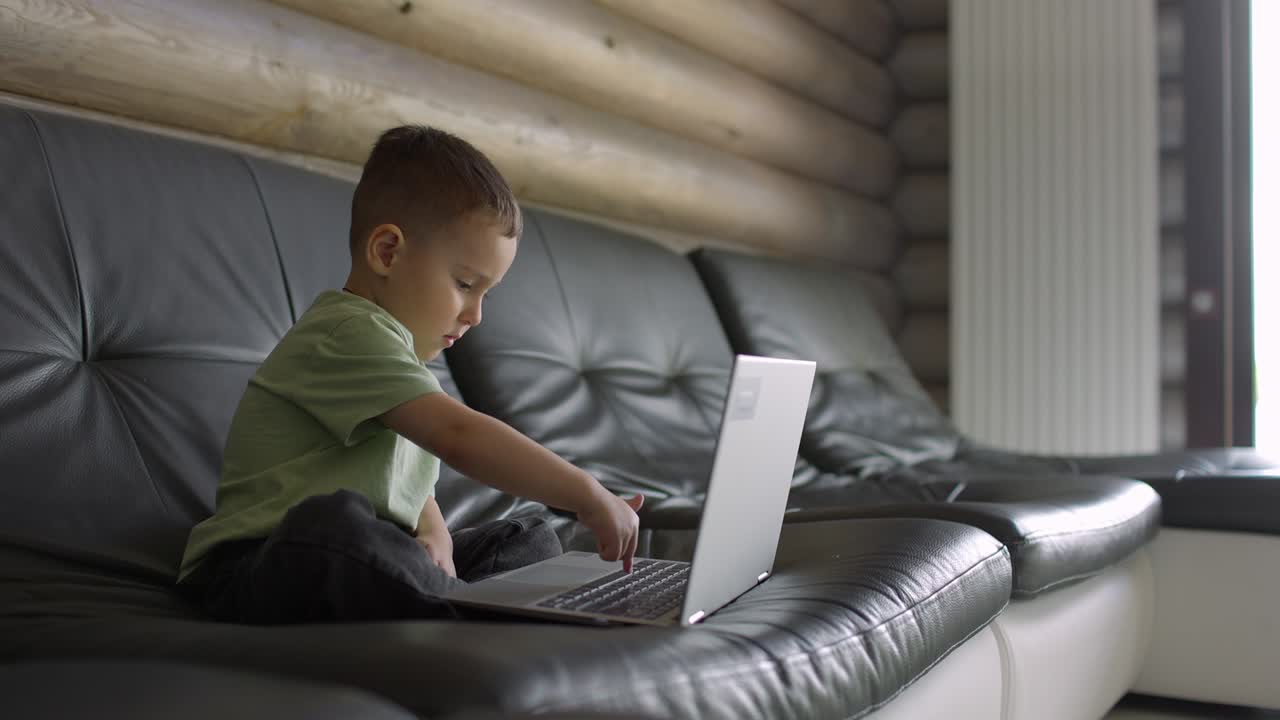 Small and cute boy learns to type on a laptop keyboard while sitting on a sofa in daylight