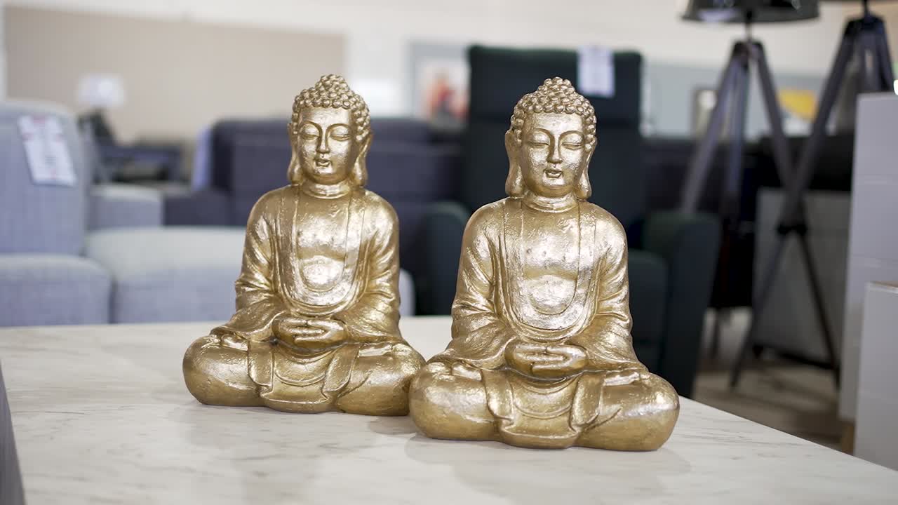 Two golden Buddha statues sit gracefully on a marble table in a bright furniture store. The serene atmosphere highlights the intricate craftsmanship and design of the statues