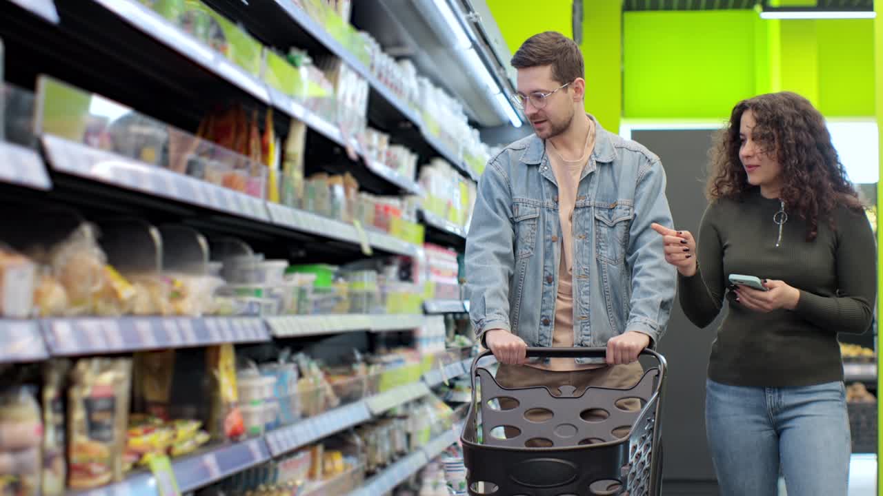 una joven pareja feliz usando un teléfono inteligente en un supermercado con un carrito de compras eligiendo productos mientras compran comestibles.