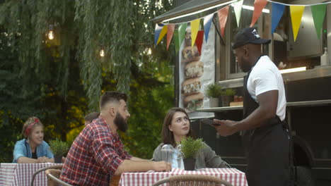 Young-Handsome-And-Cheerful-Male-Barman-In-Apron-And-Cap-Asking-For-Order-From-Happy-Couple-At-Table-Outdoor-Small-Food-Truck-Cafe-In-Park