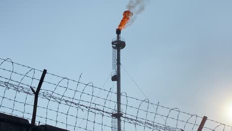 Burning-Gas-industry-flare,-refinery-industrial-area-behind-wired-wall,-tilt-up