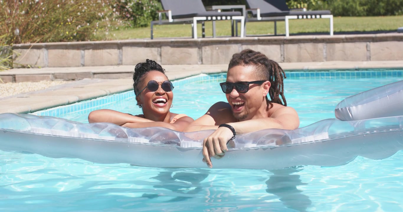 Pareja afroamericana jugando en una balsa en la piscina del patio trasero; el hombre señala, se zambulle y lleva gafas de sol