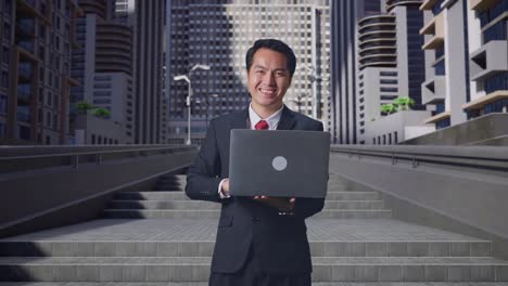an asian business man using laptop computer and smiling to camera in city