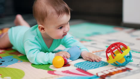 sweet little boy holds a toy in one hand and reaches for a ball with another hand. cute child lies on belly on mat playing.