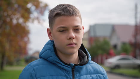 close-up of kid wearing blue jacket outdoors, looking unhappy and thoughtful, surrounded by blurred autumn trees, a red-roofed house parked car, and suburban scenery