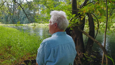 Hombre-Mayor-Se-Sienta-Junto-Al-Río,-Hierba-Alta,-árboles,-Disfrutando-De-La-Naturaleza,-Sereno