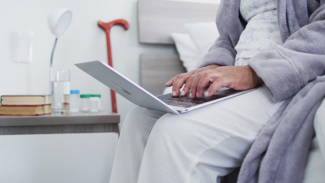 Overkrop af ældre afroamerikansk kvinde, der bruger laptop, mens hun sidder på sengen derhjemme.