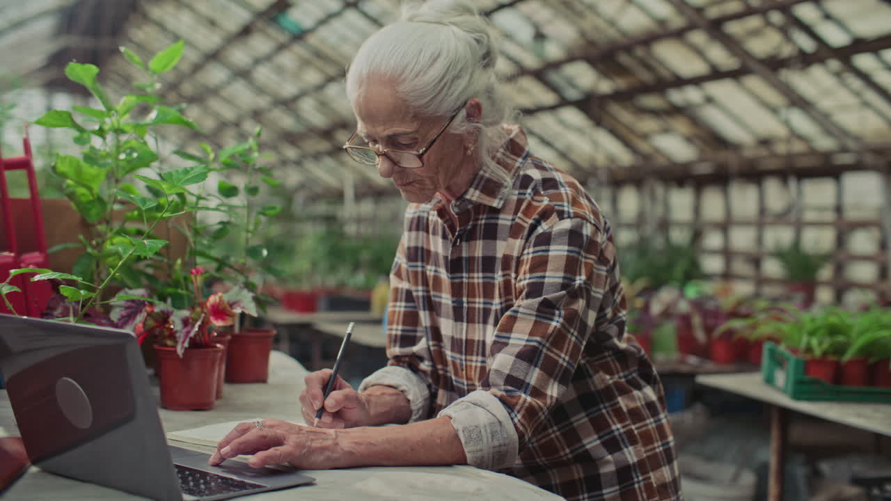 Elderly Greenhouse Manager Writing in Notebook and Using Laptop