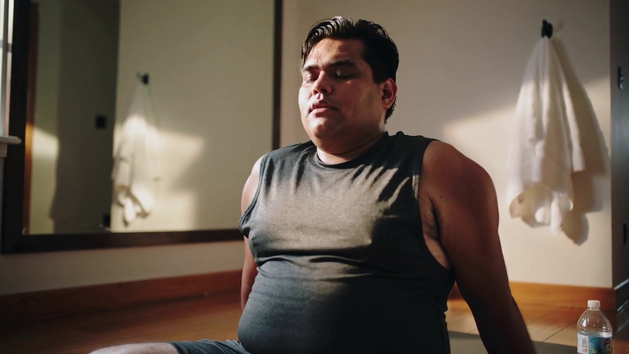 Hombre con camiseta oscura relajándose, apoyándose en las manos, cerrando los ojos y respirando en una esterilla junto a un espejo y una botella.
