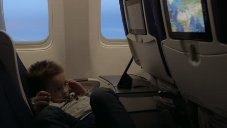 vista de un niño pequeño viendo películas en auriculares en el avión acostado en el asiento contra la ventana del avión