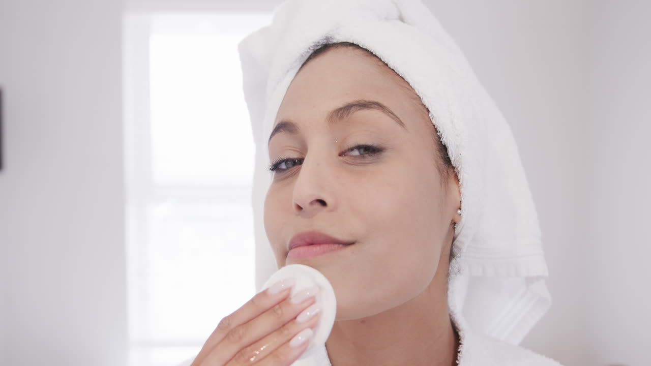 Portrait of happy biracial woman applying cleansing face using cotton pad in bathroom, slow motion