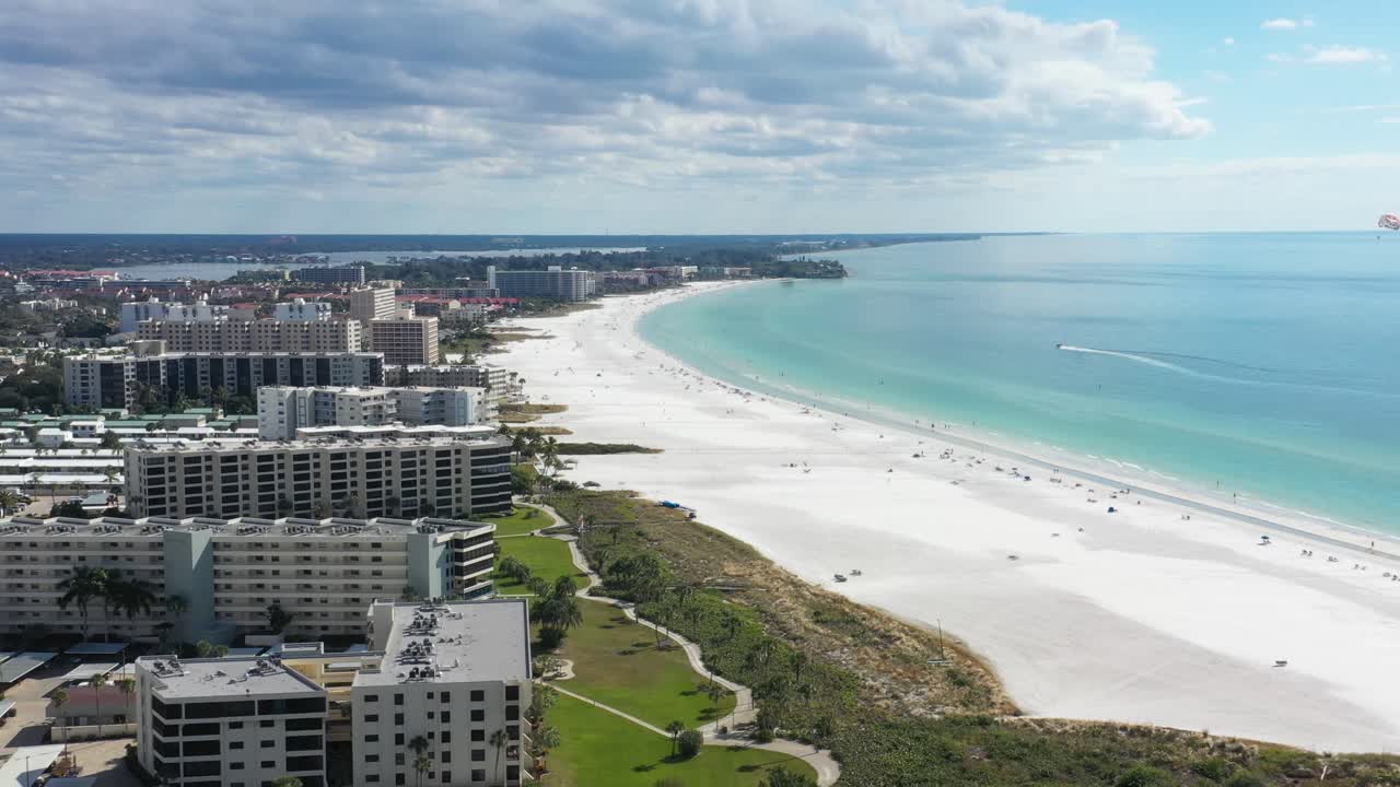 Luftoptagelse i kredsløb af den hvide sandstrand Siesta Key Beach i Florida, motorbåd sejler langs kysten, paraglider flyver over, strandlejligheder og ejerlejligheder.