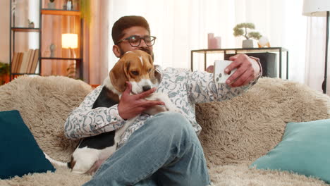 indian man watches funny dog video on smartphone while sitting on sofa with beagle licking hand