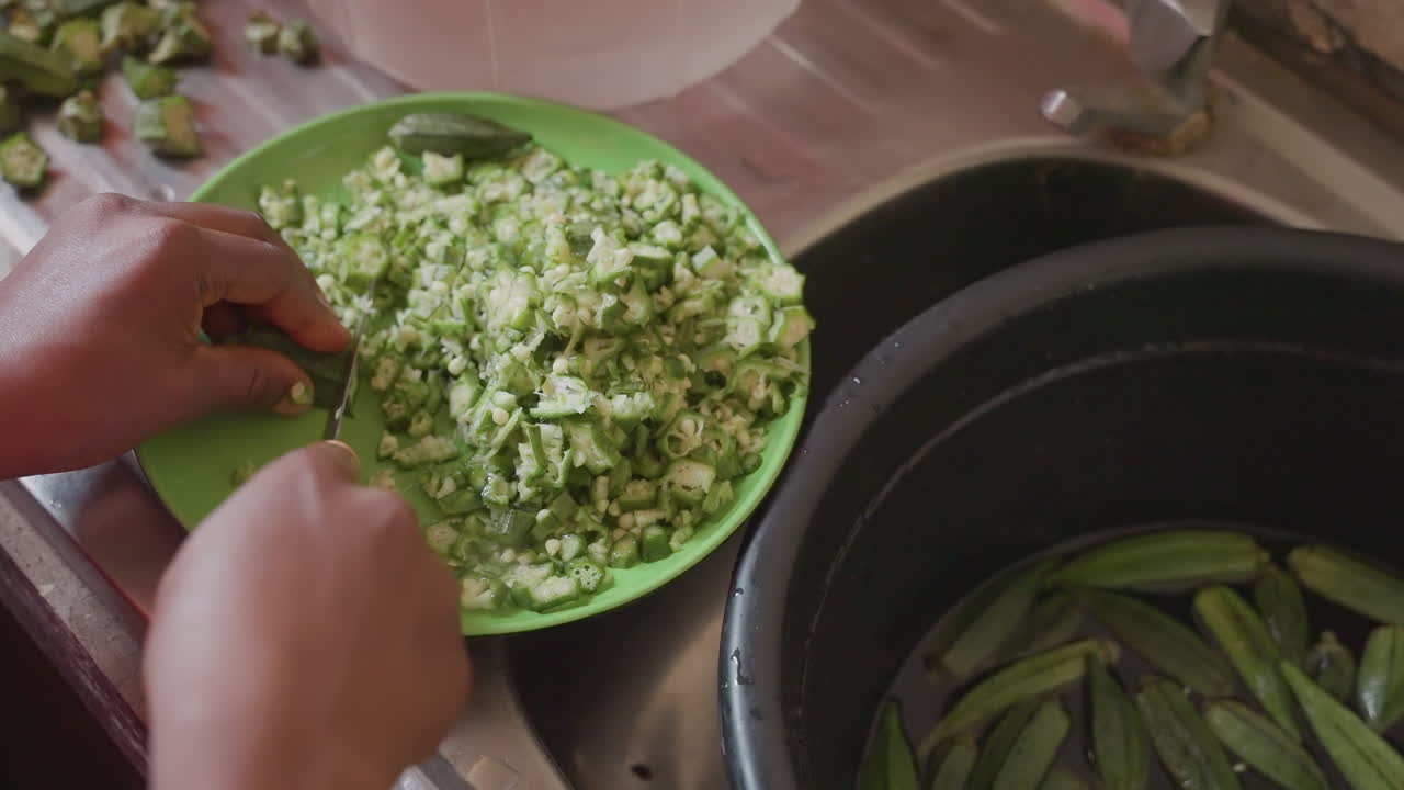 Closeup black hands transferring chopped okra into pot, communal kitchen energy, steady spoonless transfer, fresh pieces meet simmering vessel, warm family meal preparation scene