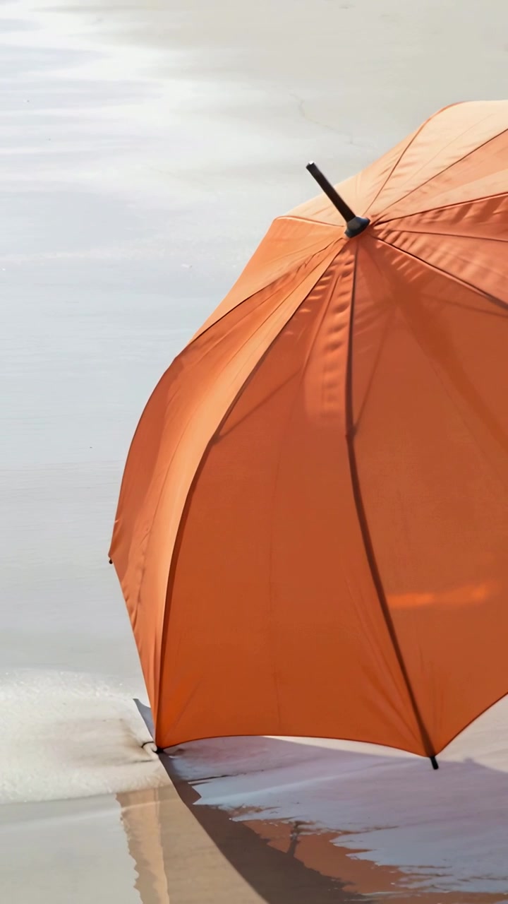 Vertikal video: Skvulpende vand bevæger en orange paraply på vådt sand, skiftende refleksion, plads til tekst.