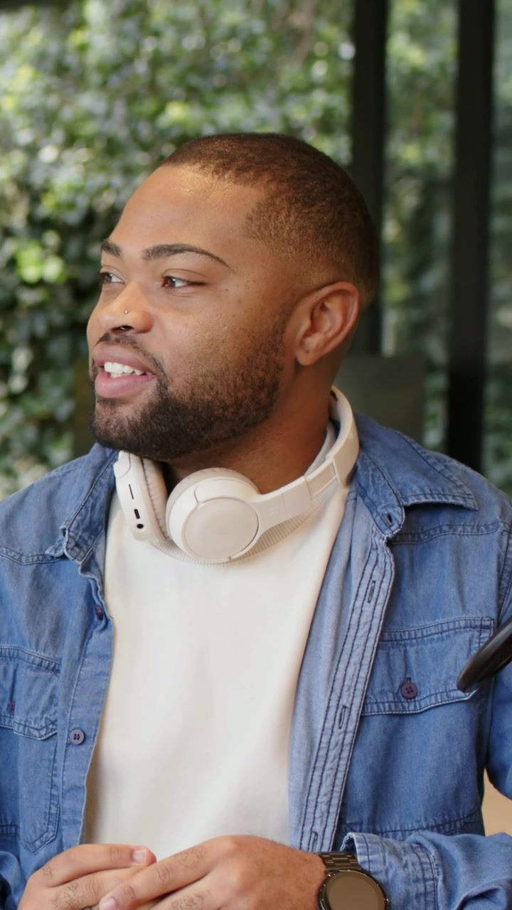 Vertical video: Talking man responding to offscreen prompt at window with headphones, denim jacket