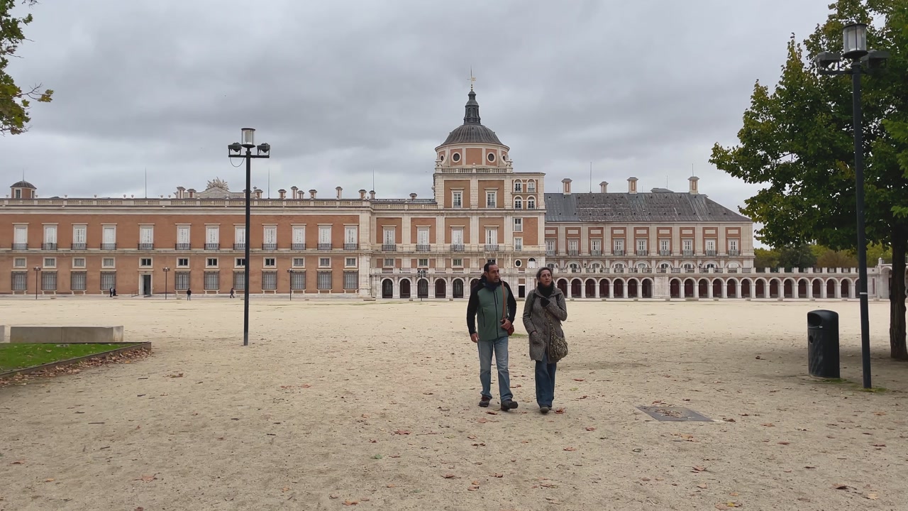 Slow-motion footage of a couple walking through the Plaza de Parejas in Aranjuez on a cloudy autumn morning, with the side of the Royal Palace in the background with its porticoes